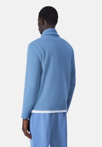 Giacca in pile azzurra chiara con colletto alto e maniche lunghe, caratterizzata da una superficie testurizzata. Abbinata a pantaloni azzurri chiari sottostanti.