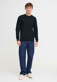Giovane uomo in piedi, indossa un maglione nero a maniche lunghe, jeans blu scuro e sneaker bianche su uno sfondo chiaro e neutro.