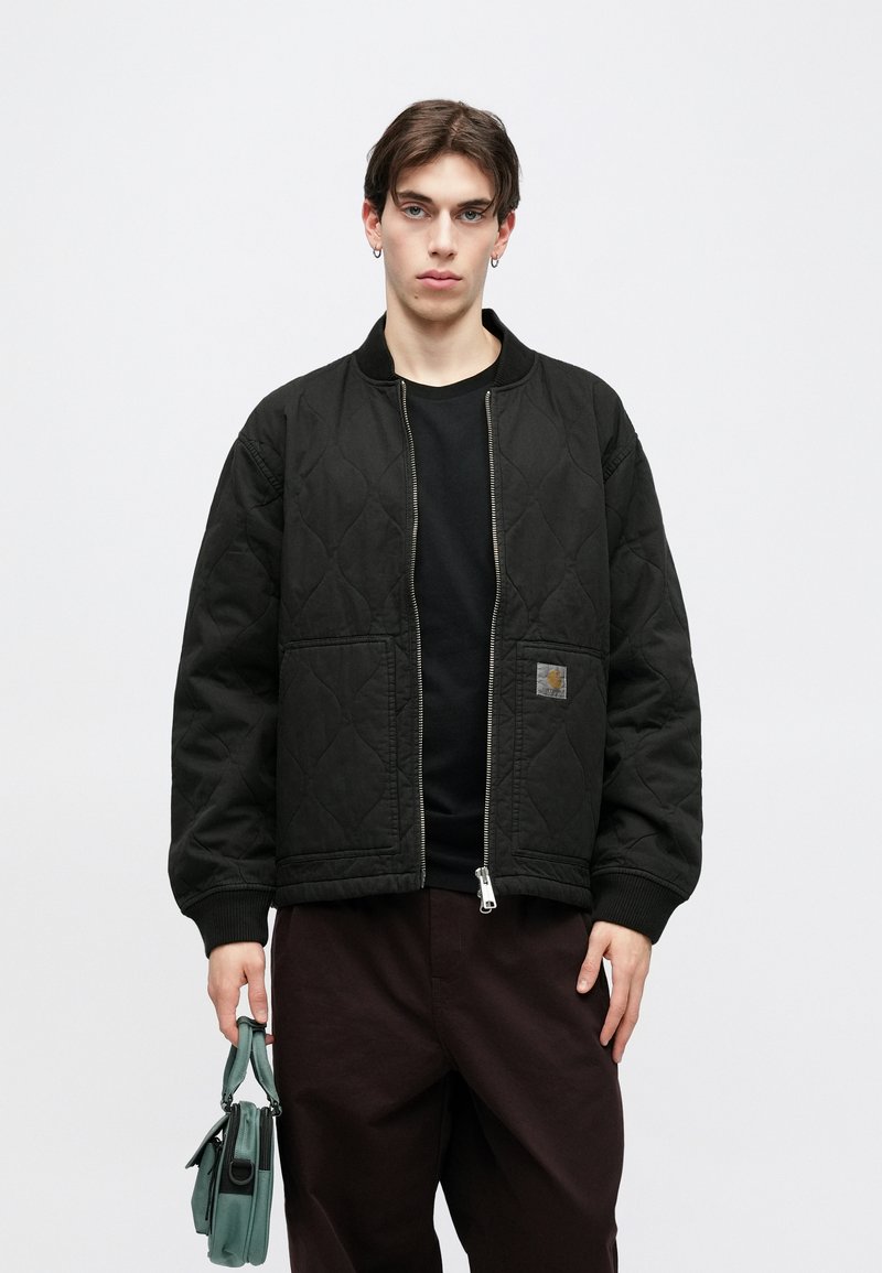 Black quilted bomber jacket with ribbed cuffs, zip closure, and side pockets. The model holds a small green handbag.