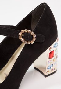 Chaussure Mary Jane en daim noir avec une bride à boucle circulaire ornée de bijoux et un talon métallique épais décoré de pierres précieuses colorées.