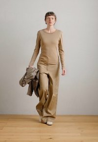 Μπέζ μπλούζα με μακριά μανίκια και χαλαρούς παντελόνους από απαλό ύφασμα, συνδυασμένη με γκρι ζακέτα ριγμένη στον ένα ώμο. Φωτεινό φόντο.