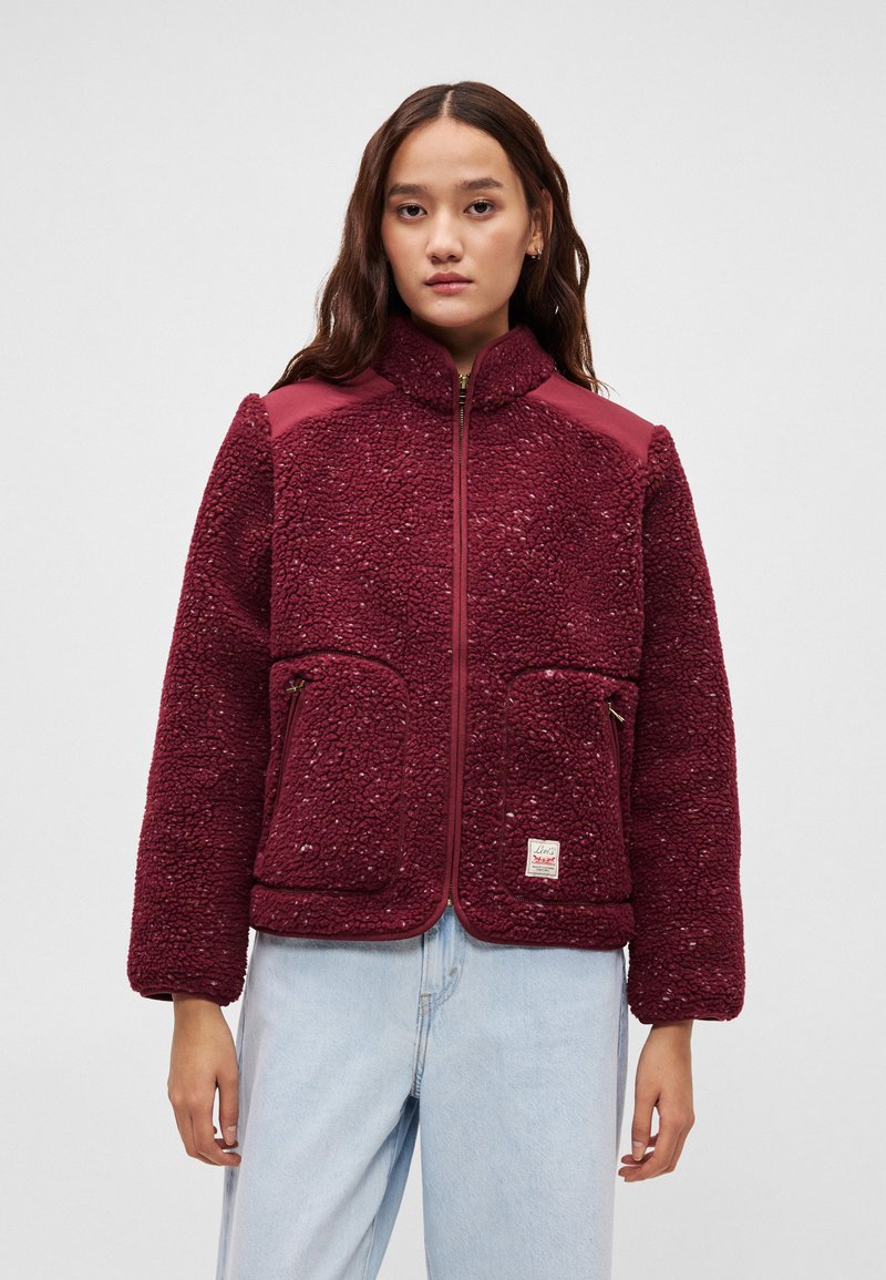 Maroon faux fur jacket with a zip front, high collar, two front pockets, and contrasting shoulder accents. Soft texture and relaxed fit.