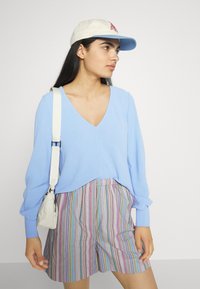 Suéter azul claro com decote em V e mangas longas, combinado com calções às riscas multicoloridos. Bolsa branca com alça de ombro. Boné bege com logótipo.