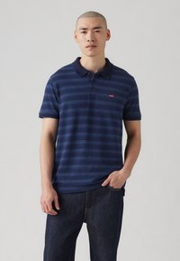 Polo de culoare albastru marin, cu dungi orizontale, mâneci scurte și guler. Are un mic logo Levi's roșu pe piept. Portat cu blugi închiși la culoare.