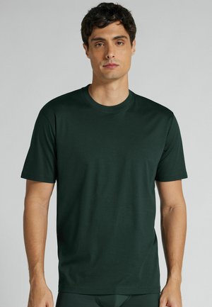 Jeune homme aux cheveux courts et foncés portant un t-shirt uni vert foncé à manches courtes, debout devant un fond gris clair.