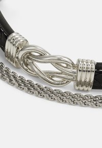 Brățară din piele neagră cu un închizător din metal argintiu împletit și o lanț metalic argintiu, evidențiind materialele și texturile contrastante.