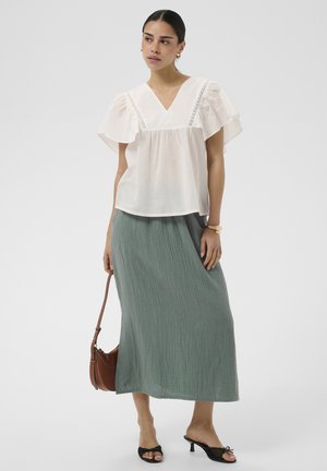 Žena nosi bielu blúzku s rozšírenými rukávmi a zelenú textúrovanú midi sukňu, drží hnedú kabelku, nosí čierne sandále na podpätku.