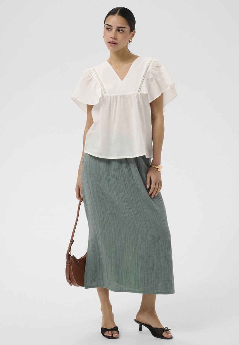 Femme portant un chemisier blanc à manches volantes et une jupe midi verte texturée, tenant un sac à main marron, portant des sandales noires à talons.