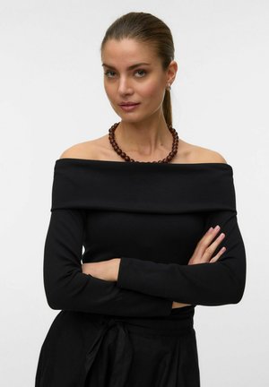 Vrouw met lichte huid en groene ogen draagt een zwart off-shoulder topje, een bruine kralenketting en heeft haar armen gekruist tegen een witte achtergrond.