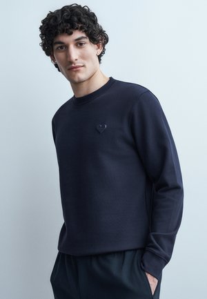 Mann mit lockigem Haar, der einen marineblauen Pullover mit einem kleinen Herz-Emblem auf der Brust trägt, vor einem hellgrauen Hintergrund stehend.