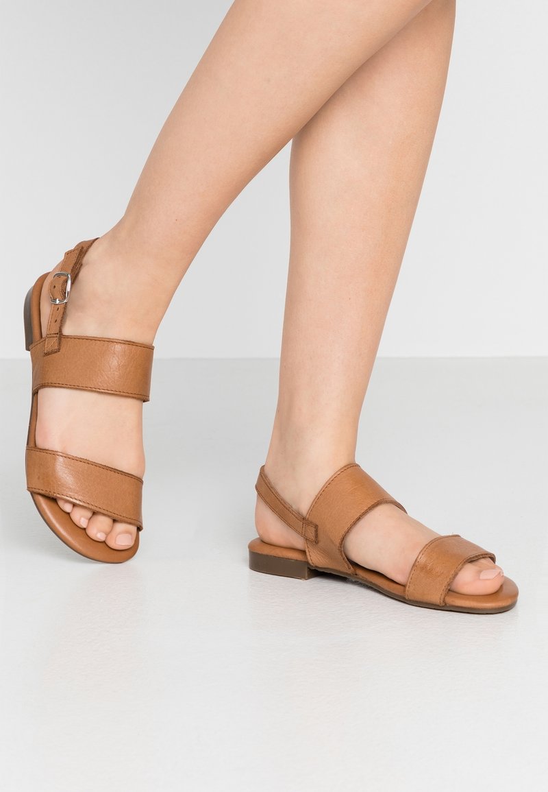 Pieds portant des sandales ouvertes en cuir marron avec des sangles à la cheville, sur un sol lisse et clair, avec un pied légèrement levé.