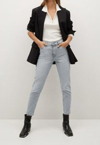 Czarna marynarka, biała bluzka z krótkim rękawem i zamkiem 1/4, jasnoniebieskie dopasowane dżinsy i czarne botki za kostkę ze kwadratowymi noskami, stojąca na neutralnym tle.