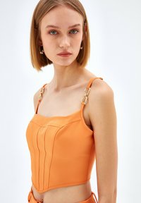 Oranssi crop top, jossa on jämäkkä muotoilu, sisältäen pystysuorat saumat ja koristeelliset kultaketjuakselit säädettävillä olkahihnoilla.