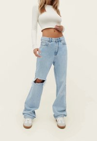 Jeans de tiro alto en azul claro con un corte suelto, que presentan una gran sección rasgada en la rodilla y un dobladillo sin terminar. Combinados con zapatillas blancas.