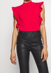 Červený volánkový top bez rukávů s vysokým límcem, v kombinaci s černými přiléhavými kalhotami z umělé kůže. Hladká textura, výrazné volánky podél ramen.
