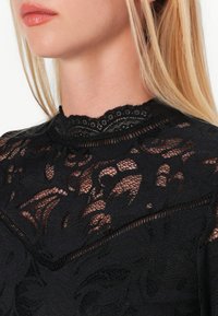 Black lace top featuring a high collar with scalloped edges, floral patterns, and sheer overlay, highlighting intricate textures and detailing.