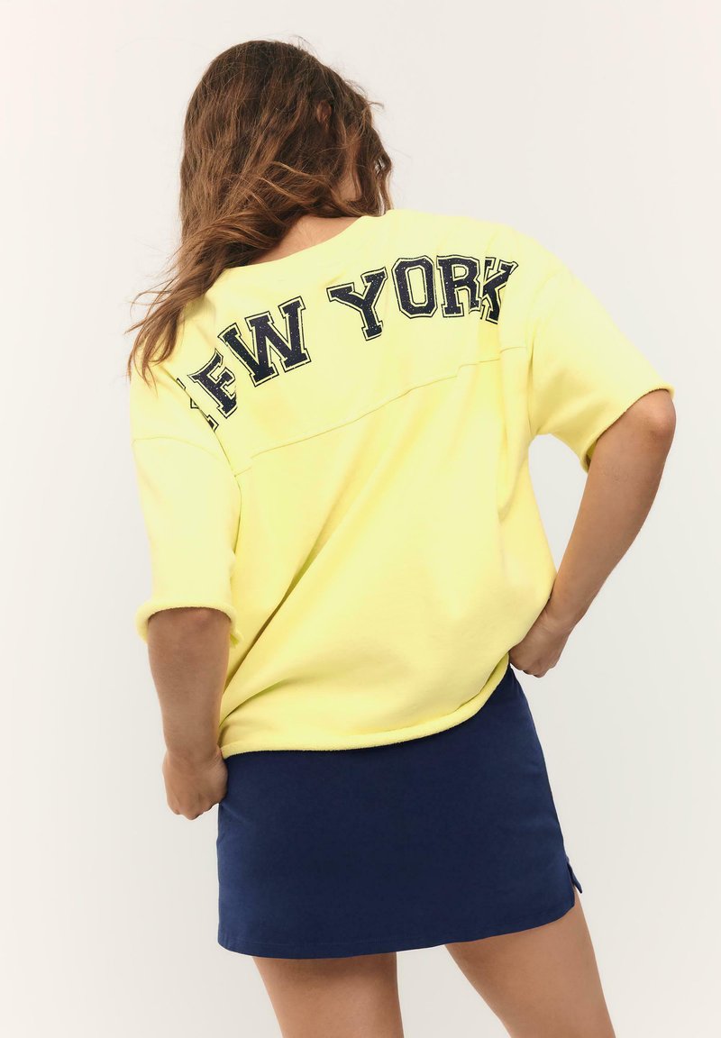 Femme aux cheveux ondulés portant un sweat-shirt jaune ample avec "NEW YORK" au dos et une jupe bleu marine, debout les mains sur les hanches.