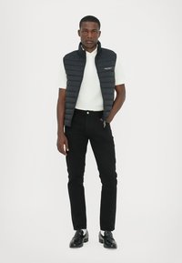 Padded schwarze Weste über einem weißen Polo, kombiniert mit schwarzen Hosen und schwarzen Schuhen. Verfügt über einen Reißverschluss und ein Logodetail auf der Weste.