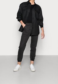 Černá košile s knoflíkovým zapínáním a náprsními kapsami, spárovaná s tmavými úzkými kalhotami a bílými teniskami. Hladká textura s přiléhavými manžetami.