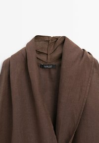 Veste en lin marron avec un large col châle et une étiquette noire indiquant "Massimo Dutti Made in Morocco" à l'intérieur de l'encolure.
