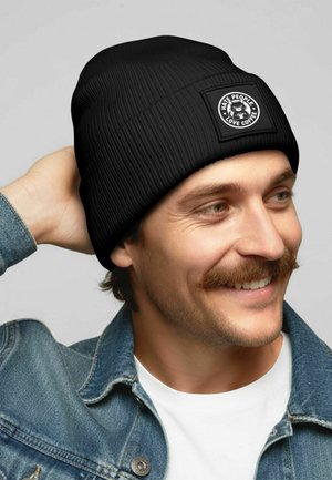 Black ribbed beanie with a square patch, featuring a graphic design in white, worn casually. Soft texture and fitted shape.