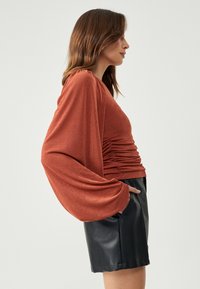 Rustrød langærmet top med en afslappet pasform, der har samling detaljer ved taljen. Sammen med tætsiddende sorte lædershorts.