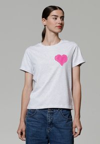 Graues Kurzarm-T-Shirt aus weicher Baumwolle mit einem rosa Herz-Graphic auf der linken Brust und einem lässigen Schnitt. Kombiniert mit blauen Jeans.