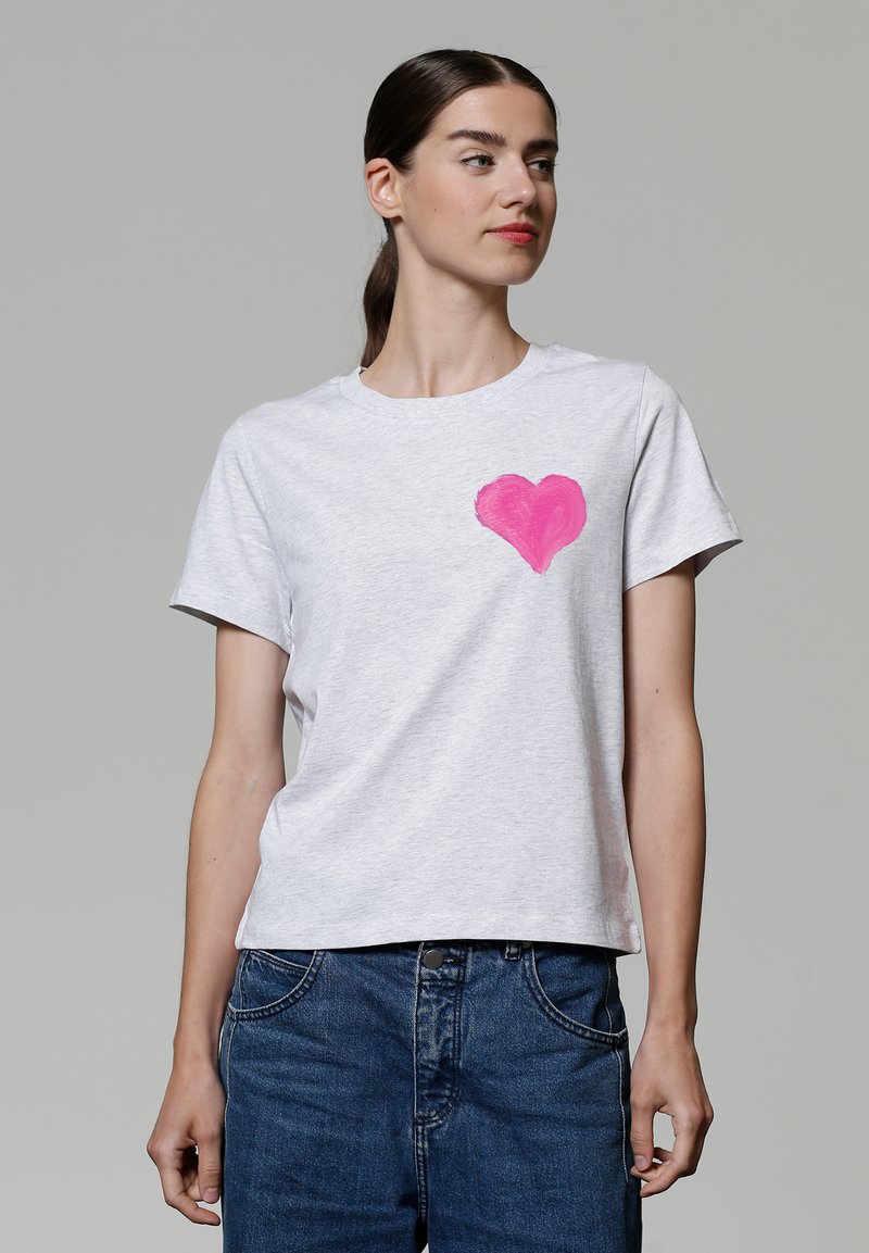 Graues Kurzarm-T-Shirt aus weicher Baumwolle mit einem rosa Herz-Graphic auf der linken Brust und einem lässigen Schnitt. Kombiniert mit blauen Jeans.