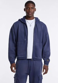 Marineblauwe zip-up hoodie gemaakt van zacht materiaal, met een trekkoordcapuchon, voren zakken en een losse pasvorm. Gedragen boven een wit T-shirt.