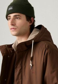 Brun ytterjacka med en beige fleece-fodrad huva, vit dragsko och en liten märkespatch. Olivgrön stickad mössa bärs på huvudet.