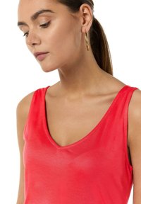 Czerwona bezrękawnikowa bluzka z dekoltem w serek, o gładkiej fakturze i lekkim materiale, uzupełniona złotymi kolczykami w kształcie kół.