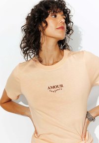 T-shirt couleur pêche en tissu doux avec un col rond. Présente le texte marron "AMOUR toujours" dans un design incurvé sur la poitrine.