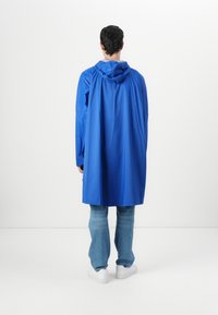 Blå, vattentät jacka med huva, långa ärmar och en lös design. Bärs över ljusa denimjeans och vita sneakers.