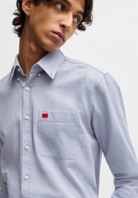 Camicia button-up azzurro chiaro realizzata in tessuto testurizzato, con taschino sul petto decorato da un distintivo rosso, e bottoni bianchi lungo la parte anteriore.
