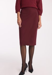 Burgundowa spódnica ołówkowa z elastycznego materiału, charakteryzująca się gładką fakturą, o długości midi, zestawiona z czarnymi szpilkami.