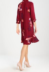 Burgunderfarbenes Blumen-Kleid mit Dreiviertelärmeln, gerafftem Saum und floralen Mustern in Pink und Gelb. Trägt man mit nuden High-Heels.