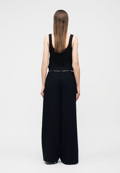 Γυναίκα με ίσια μαλλιά, που φοράει μαύρο αμάνικο τοπ και μαύρο παντελόνι ψηλόμεσο με φαρδιά πόδια, στέκεται με την πλάτη προς τα εμπρός σε απλό φόντο.