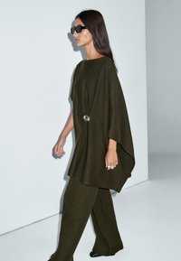 Donna con capelli lunghi e scuri indossa una maglia verde scuro larga con spilla e pantaloni a gamba larga abbinati, camminando contro uno sfondo bianco uniforme.