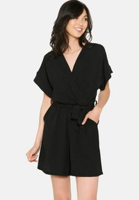 Czarny kombinezon z krótkim rękawem, dekoltem w serek i paskiem w talii. Wykonany z lekkiego materiału, o luźnym kroju i bocznych kieszeniach.