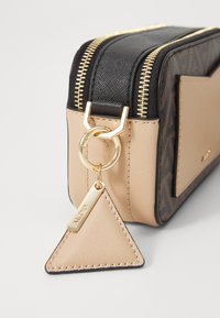 Sac structuré crème et noir avec fermeture éclair dorée, arborant une étiquette triangulaire et des surfaces texturées. Les accents comprennent un matériel en anneau à pression.