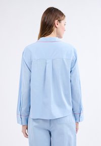 Světle modrá košile s dlouhým rukávem a límcem, s červeným lemováním podél okrajů a detailním skladem na zádech. Hladká textura látky.