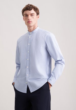 Młody mężczyzna w jasnobłękitnej i białej koszuli w paski z kołnierzem typu mandarynka i guzikami, z rękami w kieszeniach, na tle jasnego, gładkiego tła.