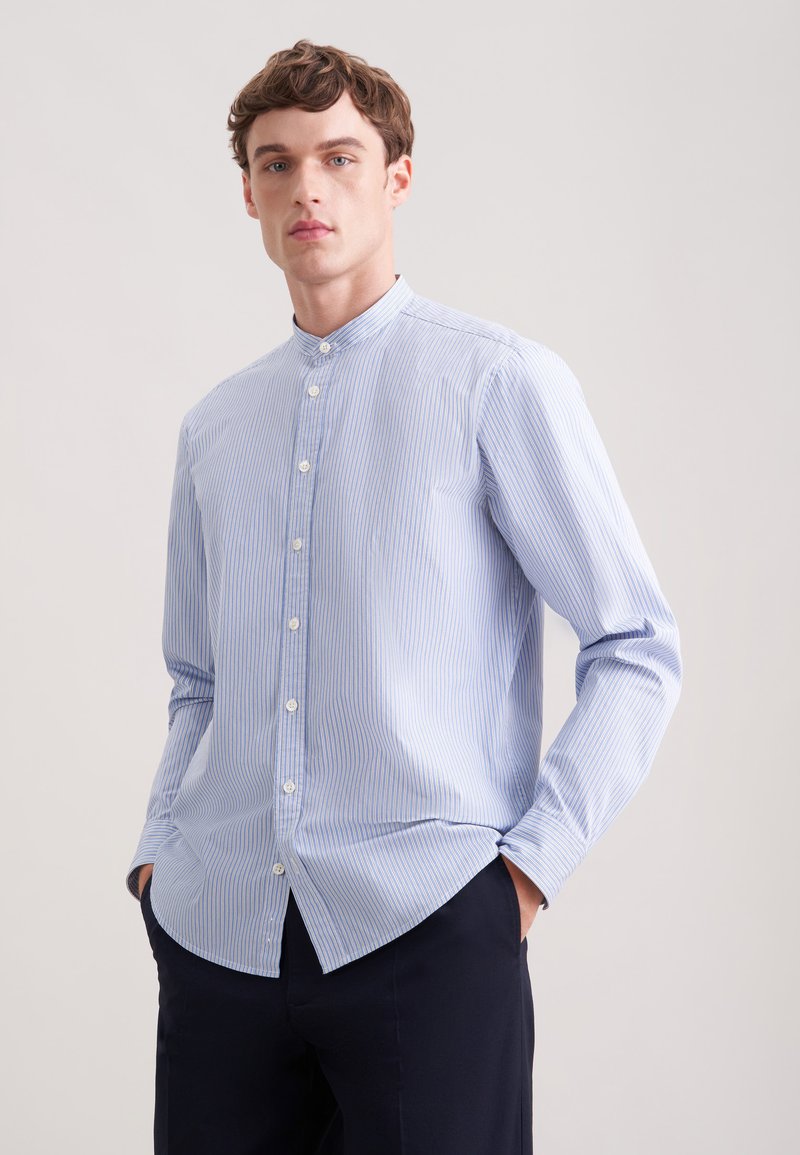 Jeune homme portant une chemise à col mandarin à rayures bleu clair et blanches avec boutons, les mains dans les poches, devant un fond clair uni.