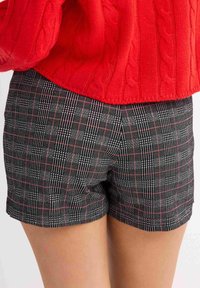Pantalones cortos de cuadros en blanco y negro con detalles en rojo, que presentan un diseño a medida y una textura suave, combinados con un suéter de punto trenzado rojo.