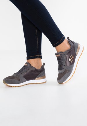 Osoba nosząca ciemnoszare i brązowe buty sportowe Skechers z białymi podeszwami i gumowym bieżnikiem, w połączeniu z ciemnoniebieskimi obcisłymi dżinsami.