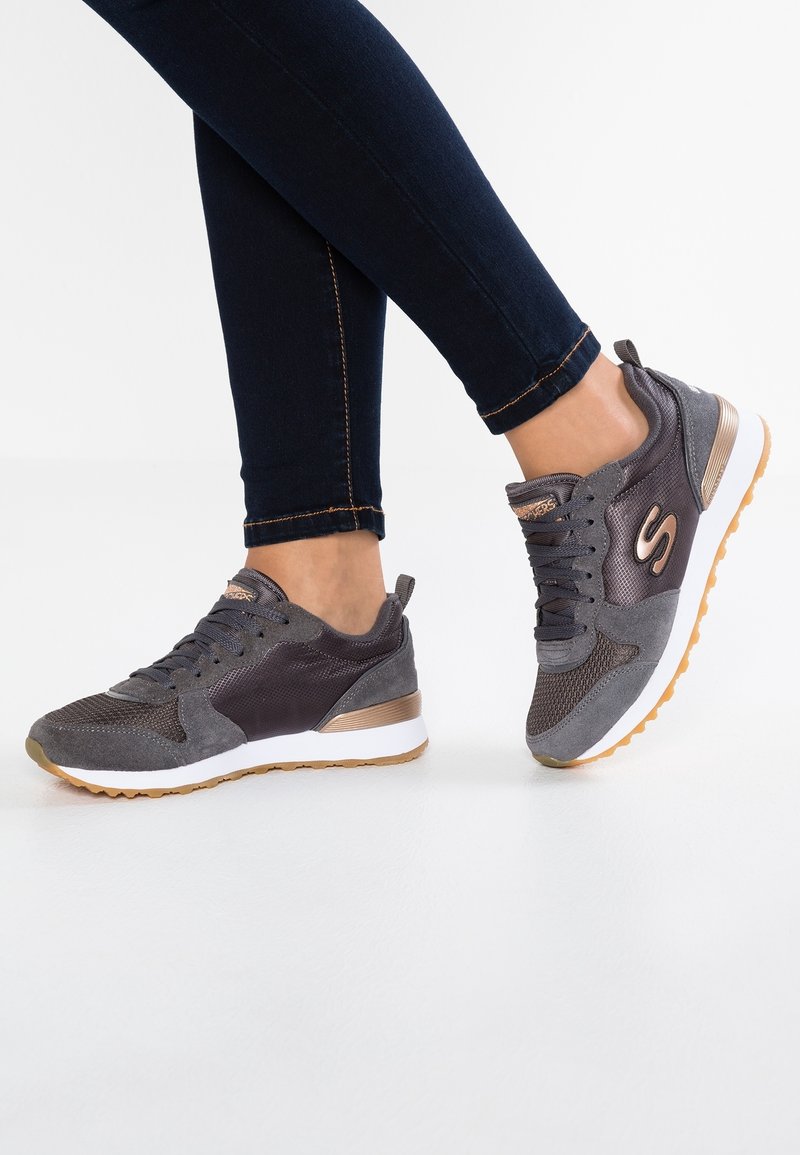 Personne portant des baskets Skechers gris foncé et bronze avec des semelles blanches et des crampons en gomme, associées à un jean skinny bleu foncé.