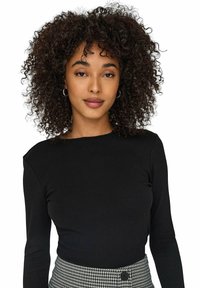 Maglietta nera a maniche lunghe con scollatura rotonda abbinata a una gonna a scozzese con dettaglio di bottone in vita. Capelli ricci.