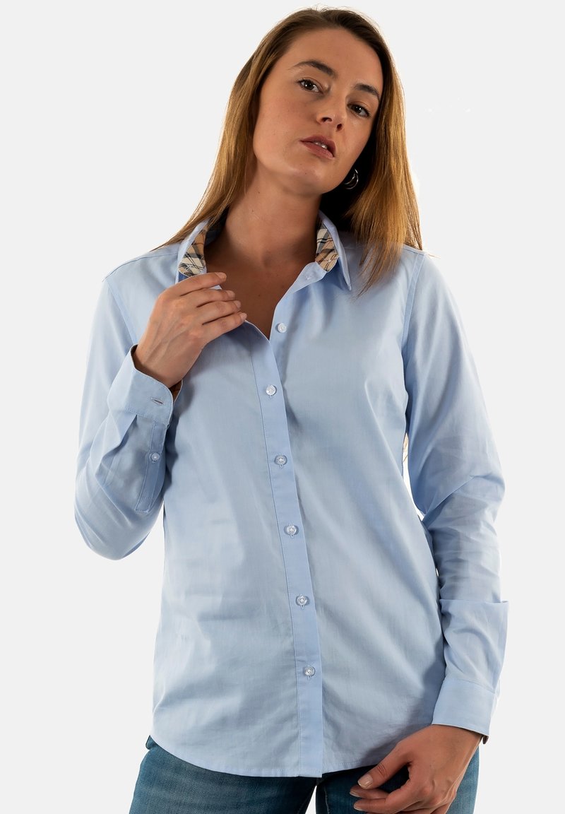Vrouw die de kraag van een lichtblauwe overhemd met patroon aan de binnenkant aanpast, gekleed in een spijkerbroek tegen een effen achtergrond.