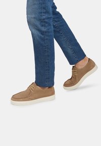 Pantofi din suede maro cu talpă din cauciuc crem �și detalii de șiret, purtați cu blugi, arătând un design casual și discret.