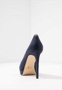 Pump con tacco alto di colore blu navy, realizzato in materiale testurizzato, con punta affusolata e logo Michael Kors sul tacco e sulla suola, esposto su una superficie bianca.